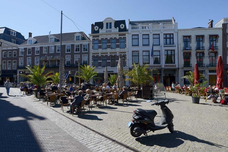Leute sitzen in einem Gastgarten auf einem verkehrsberuhigten Platz im Zentrum von Den Haag, abgestellter Motorroller, Häuserzeile, Palmen