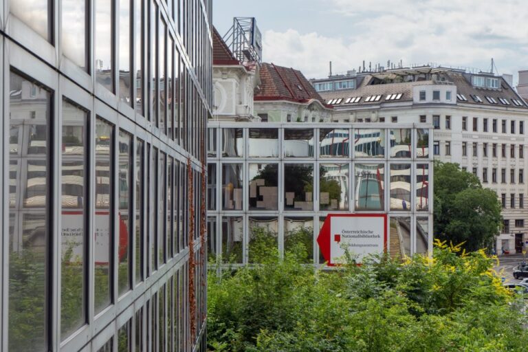 Gebäude mit Glasfassade, dahinter Gründerzeithäuser, Büsche, Baum