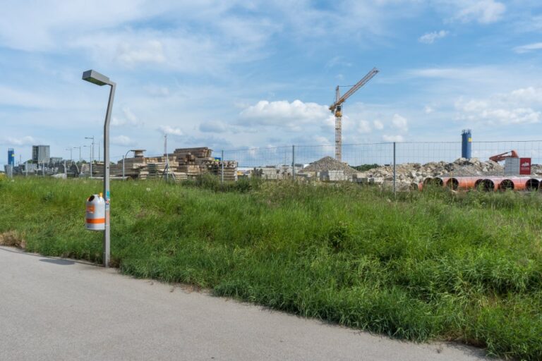 Gehsteig, Laterne und Müllkübel neben der Baustelle der Stadtstraße, Kran, Zaun, Grünstreifen