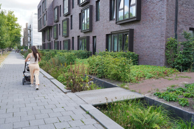 Begrünung in Amsterdam, Gebäude mit großen Fenstern und Klinkerfassade, Frau schiebt einen Kinderwagen