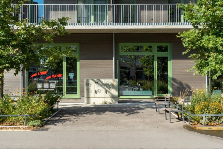 Schaltkasten vor einem Haus mit Holzfassade, Sessel, Grünflächen, Bäume, Schaufenster