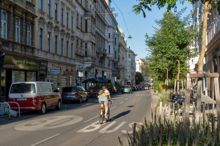 Radfahrer und Autos fahren durch die Burggasse, rechts Bäume