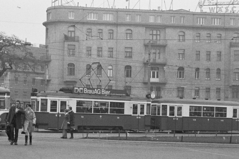 Straßenbahn und Leute vor einem Jahrhundertwendehaus mit vereinfachter Fassade